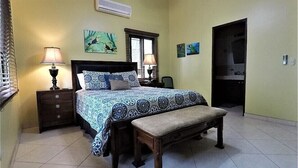 Una camera da letto con un letto, due comodini con lampade, una panca, una finestra con persiane e opere d'arte alle pareti.