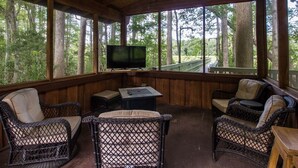 A covered outdoor seating area with wicker furniture, a television, and a wooden deck overlooking a forest. - Secluded Waterfront Retreat With Private Dock                                    (Ocklawaha)