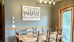Un espace repas avec une table et des chaises en bois, un panneau fixé au mur et une fenêtre avec des stores.