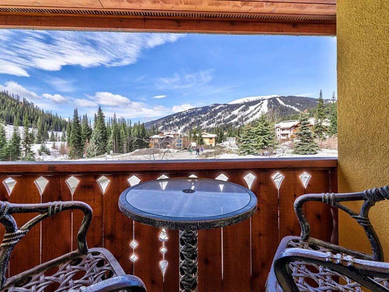 A balcony with a round table and two wicker chairs, overlooking a mountainous landscape with snow-capped peaks and a clear blue sky.