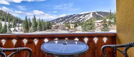 A balcony with a round table and two wicker chairs, overlooking a mountainous landscape with snow-capped peaks and a clear blue sky.
