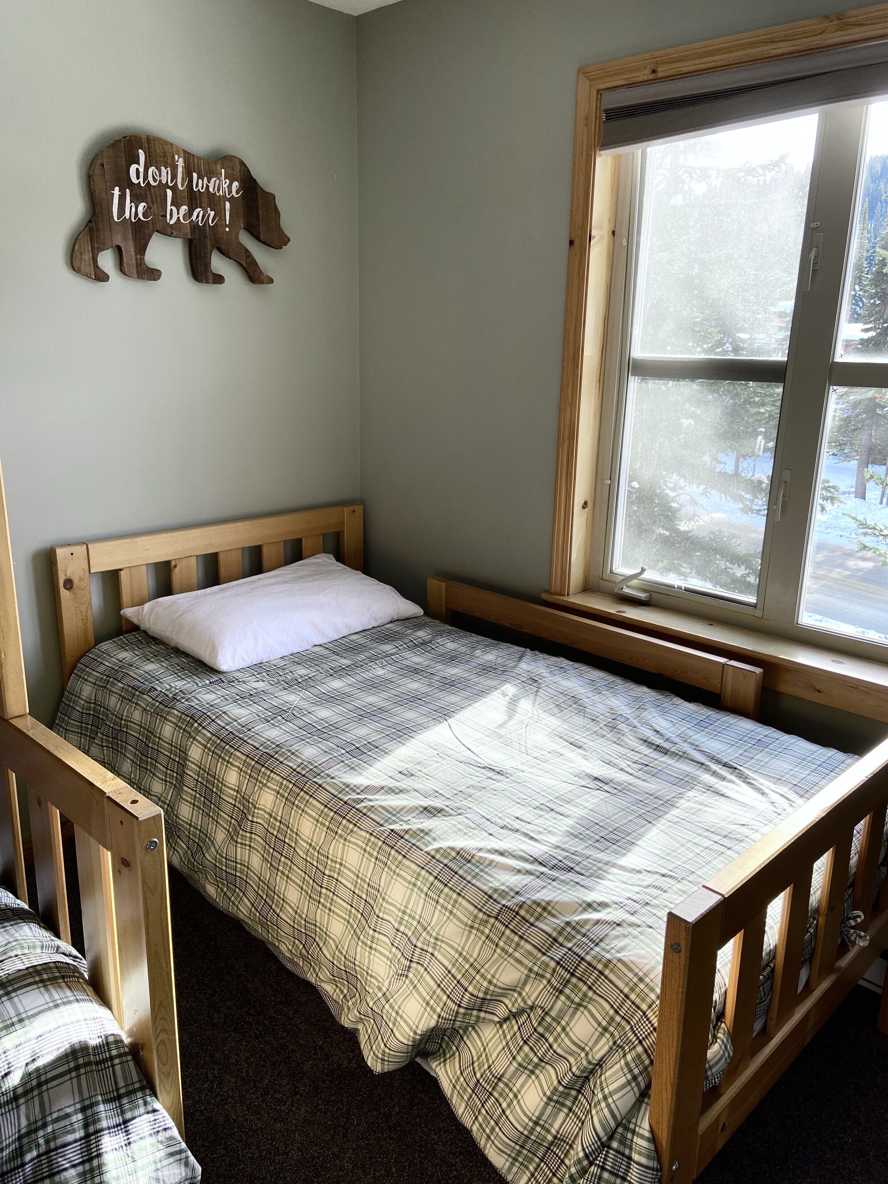 Una cama individual con colcha escocesa, cabecera de madera y ventana con vista a los árboles.