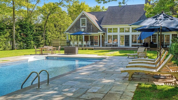 A swimming pool with a stone deck, surrounded by lounge chairs and umbrellas, with a house in the background. - Wainscott Southampton Estate Timeless luxury with a private pool,... (Wainscott)