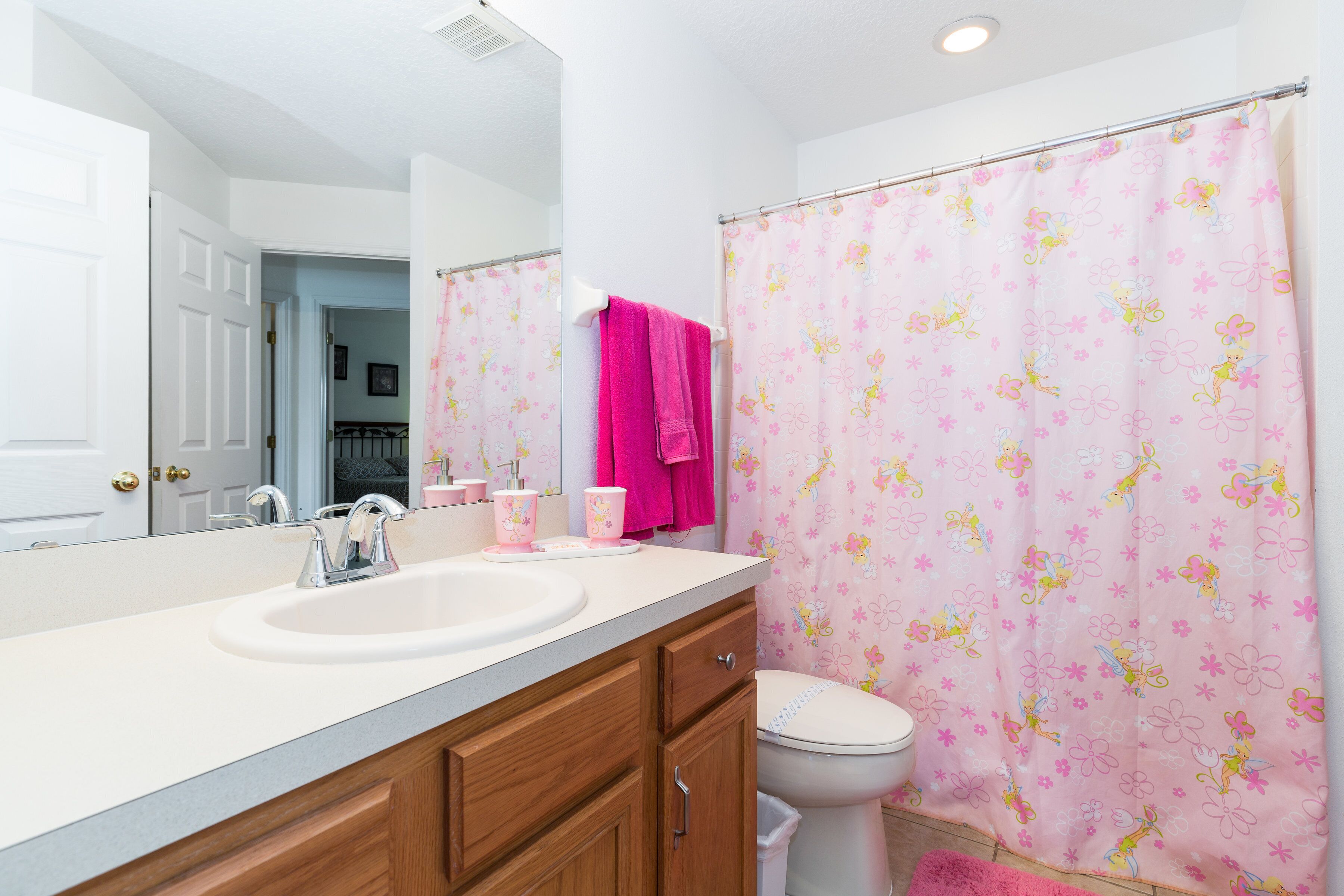 A bathroom with a white sink, a mirror, a pink shower curtain with fairy designs, and a white toilet.