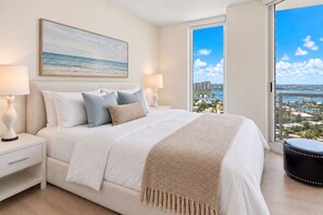 A bedroom with a bed, nightstands, a window with a city view, and a painting on the wall.