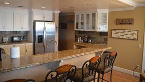 A kitchen with white cabinets, a stainless steel refrigerator, granite countertops, and a bar area with chairs. - Major's Toy Box - Amazing Home Havasu Landing  (Havasu Lake)