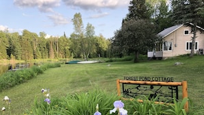 A sign reading "Forest Pond Cottage" in a grassy area with a pond, boats, and trees in the background. - Adirondack Cottage - Four Season, Pet-Friendly, Cozy & Beautiful. Read  reviews! (Lowville)