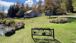 A sign for Forest Pond Cottage with a scenic view of a house, trees, and a pond. - Adirondack Cottage - Four Season, Pet-Friendly, Cozy & Beautiful. Read  reviews! (Lowville)