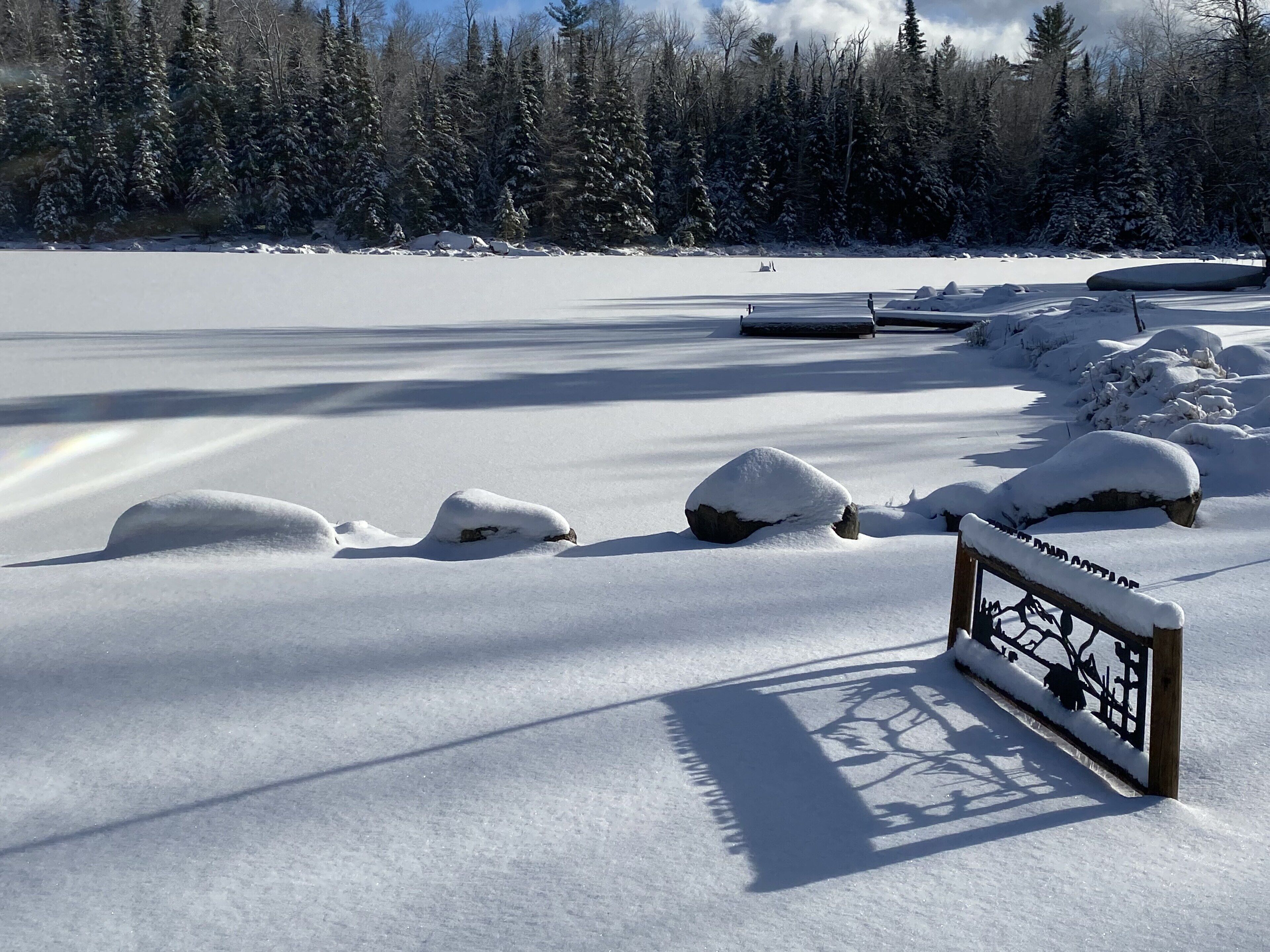 Un paysage enneigé avec un banc en bois et un panneau, entouré d’arbres et d’un lac gelé.
