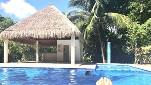 Ein Poolbereich mit einer strohgedeckten Struktur, einem klaren blauen Pool und einer Person, die auf einer Liege entspannt.