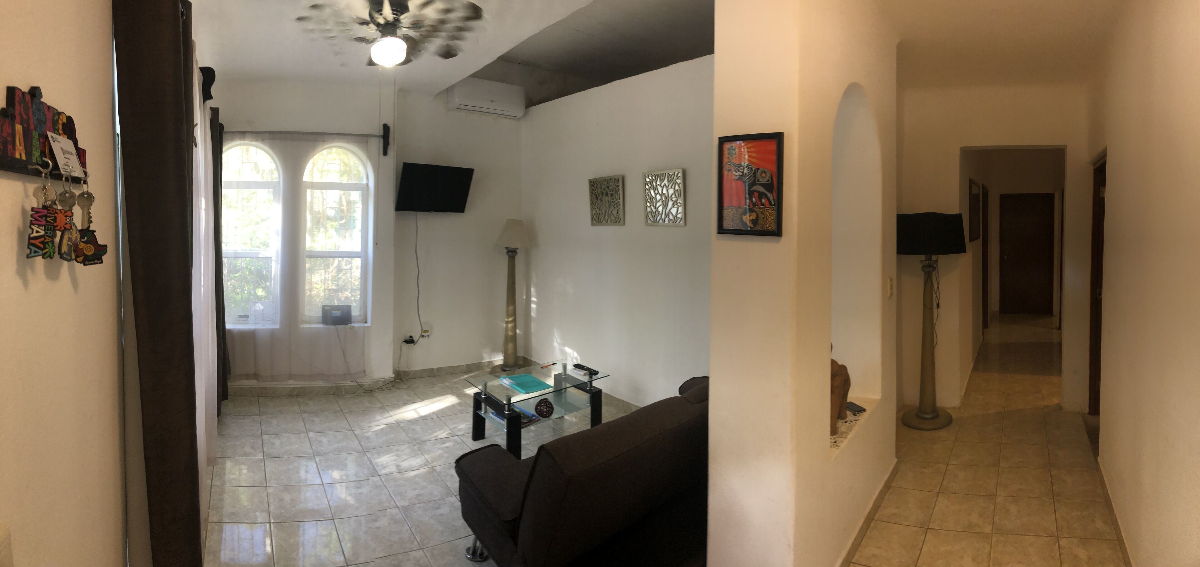 A living room with a glass coffee table, a sofa, a TV mounted on the wall, and artwork on the walls.