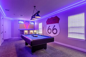 Ein Billardtisch in einem Raum mit violetter Beleuchtung, ein Neon-Schild mit der Aufschrift 'Route 66' und ein Billardständer.
