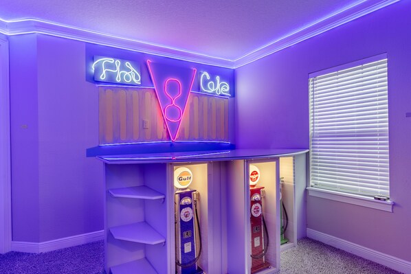 Ein Zimmer mit schräg stehender Decke, mit Neonreklame für 'Flu' und 'Coke', sowie zwei Zapfsäulen mit der Aufschrift 'Gulf'.