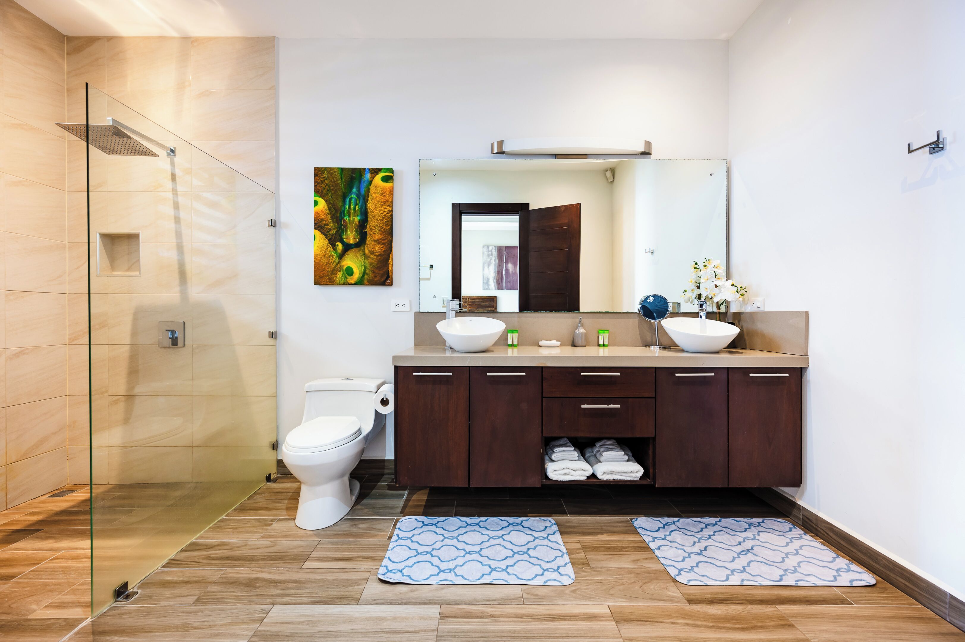 A modern bathroom with a glass-enclosed shower, a white toilet, a wooden vanity with a large mirror, and two patterned rugs on the floor.