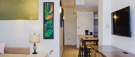 A modern living room with a sofa, a small table with a lamp, and a kitchen area with a dining table and bar stools.