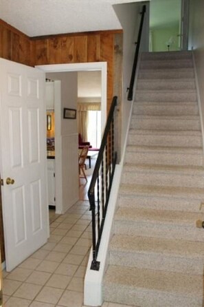 A staircase with a handrail leading to a landing, a door on the left, and a view into a room with a chair and table.