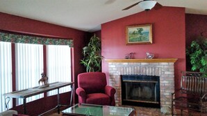 A living room with a red wall, a fireplace, a red armchair, a glass coffee table, and a window with blinds.