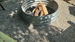 A metal fire pit with wood logs, surrounded by gravel, with two Adirondack chairs and a yellow chair in the background. - Pier Joy: Two private cottages on 2.5 wooded acres, close to Union Pier beaches (Union Pier)