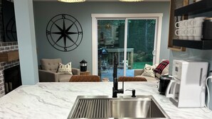 A modern kitchen with a marble countertop, stainless steel sink, and white cabinets. There are two pendant lights, a compass wall decoration, and a view of the outdoors through a sliding glass door. - Pier Joy: Two private cottages on 2.5 wooded acres, close to Union Pier beaches (Union Pier)