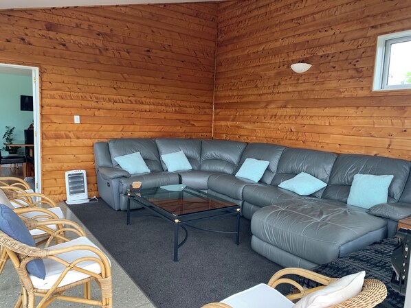 Ein gemütliches Wohnzimmer mit einem grauen Ledersofa, Korbstühlen, einem Glastisch und Holzwänden.
