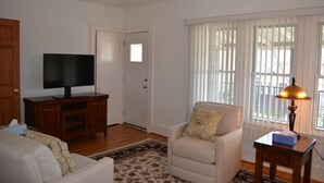 A living room with a television on a wooden cabinet, a white armchair, a small table with a lamp, and a window with blinds. - HomeAway Residential Apartment (Dearborn)