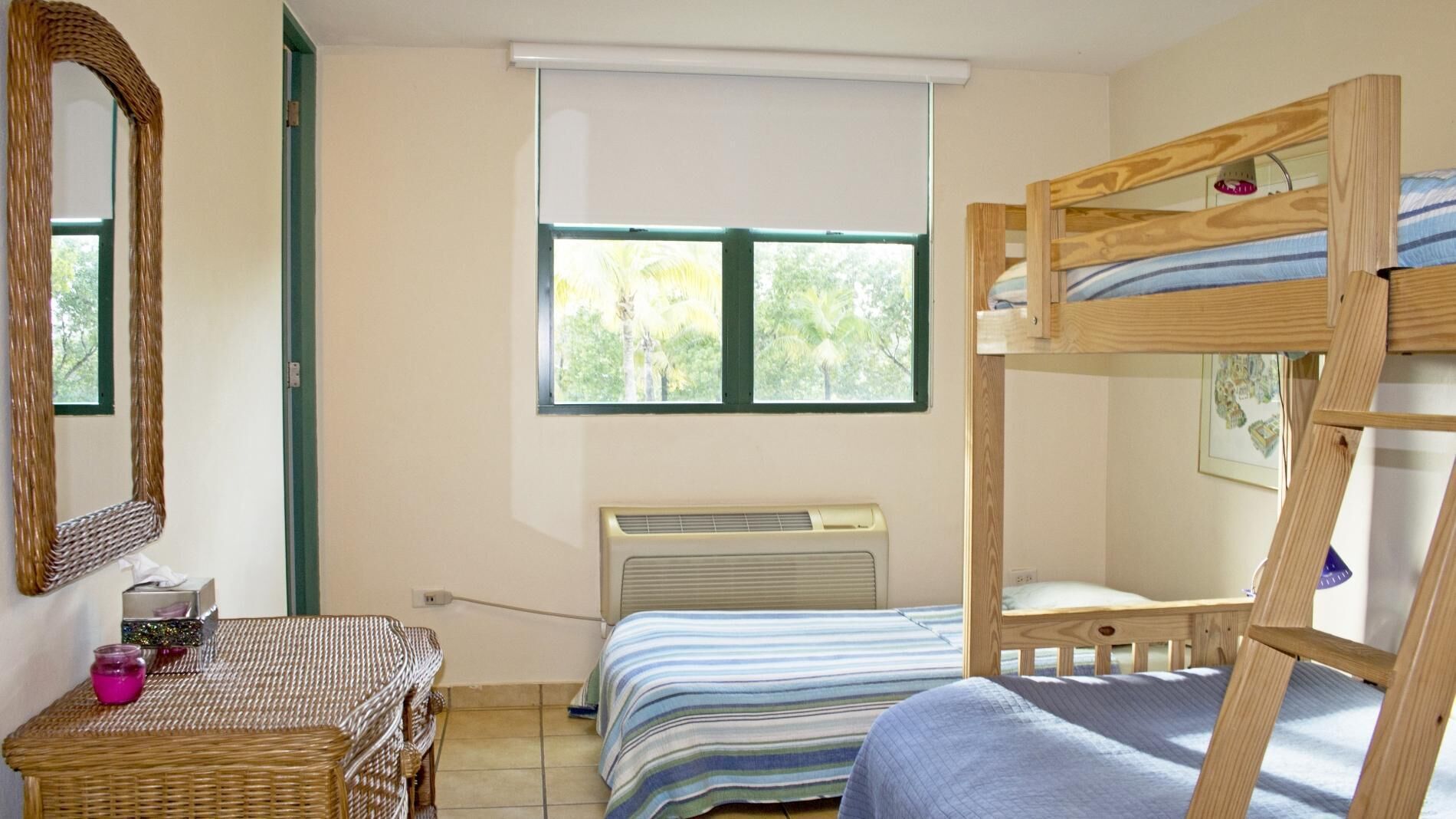 A room with a bunk bed, a desk with a chair, a large mirror, and a window with a view of greenery.