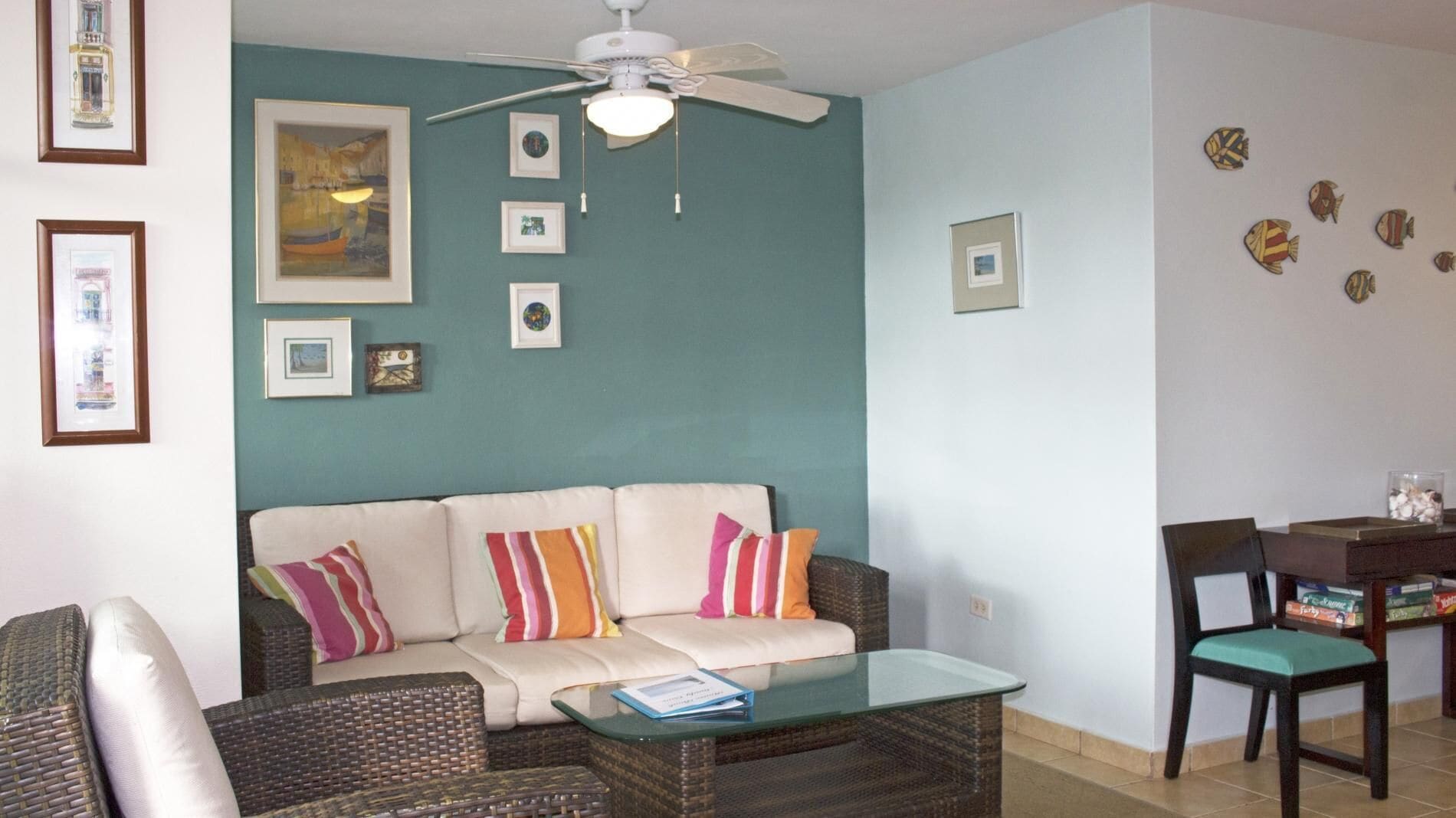 A living room with a sofa, coffee table, and a ceiling fan. There are framed pictures on the walls and a small table with a chair.