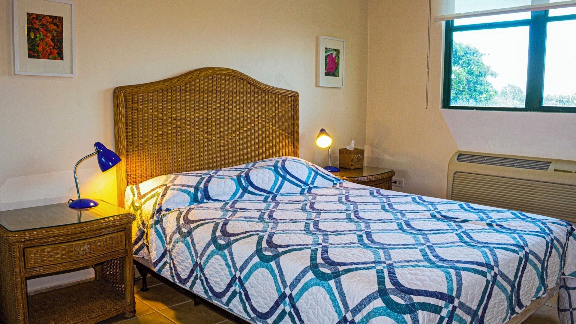 Une chambre à coucher avec une couvre-lit à motifs, une tête de lit en osier, une lampe de chevet et une fenêtre donnant sur des arbres.