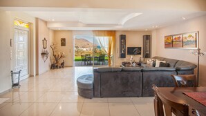 A spacious living area with a large gray sofa, a dining table, and a view of a mountainous landscape through large glass doors. - Villa Balos with fantastic sea view (Chania)