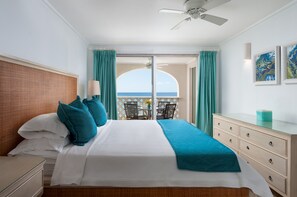 Ein Hotelzimmer mit Bett, Kommode, Nachttisch, Schreibtisch mit Stühlen und Blick auf das Meer durch eine Glastür.