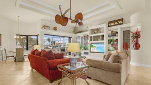 Un ampio soggiorno con un divano rosso, un divano beige e un tavolino in vetro. Si nota una ventola a soffitto, un lampadario e una grande finestra con vista su palme.