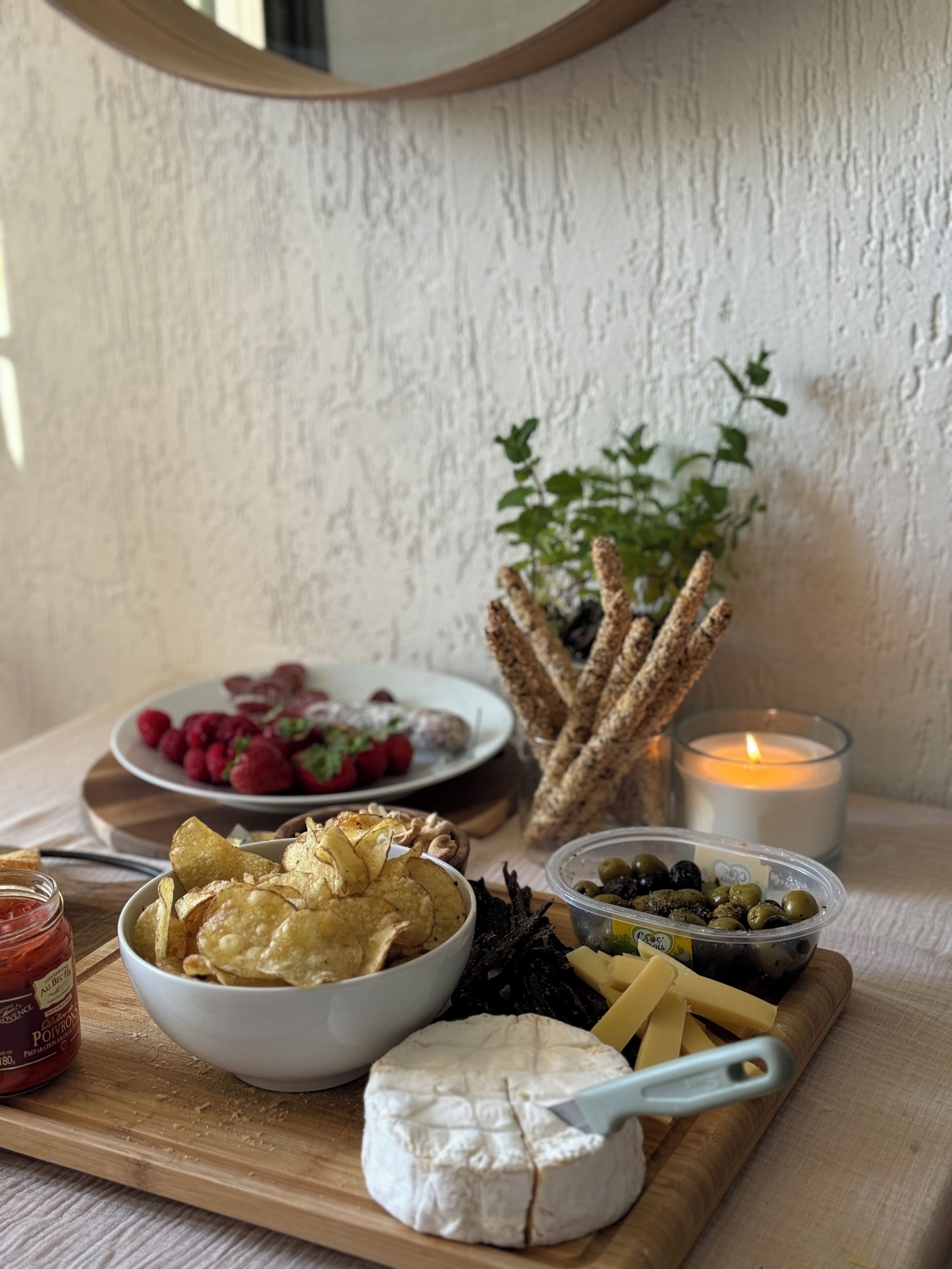 A wooden board with various snacks and cheeses, a candle, and a potted plant.
