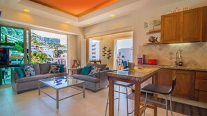 Ein modernes Wohnzimmer mit Sofa, Couchtisch und Barhocker. Eine Küche mit Holzausstattung und Blick auf die Stadt ist durch die Glastür sichtbar.