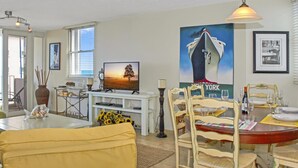 Un coin repas avec une table ronde dressée pour quatre personnes, un téléviseur diffusant une scène de coucher de soleil et une affiche de navire accrochée au mur.