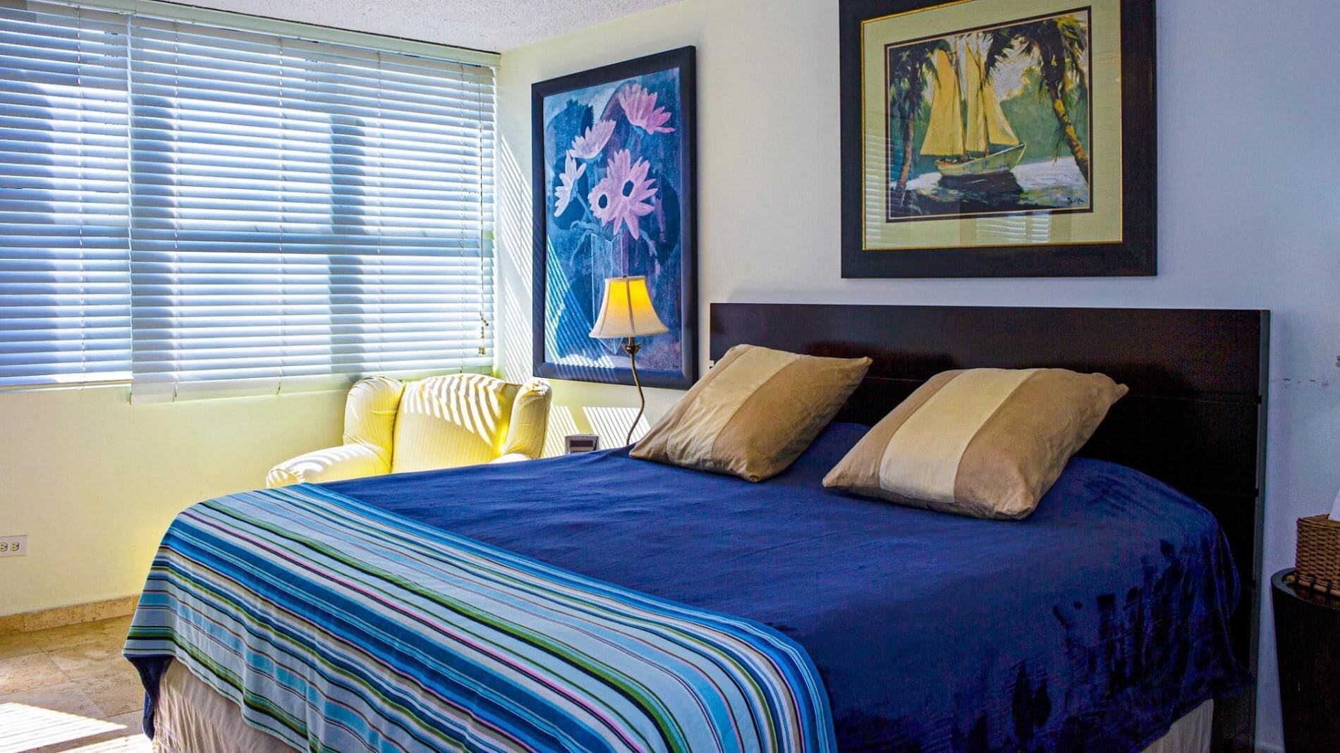 A bedroom with a large bed, a window with blinds, and two framed artworks on the wall.
