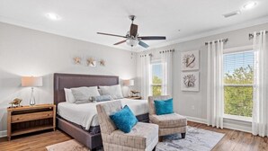 Une chambre à coucher comprenant un lit, deux fauteuils, une table de chevet, un ventilateur de plafond et une grande fenêtre avec des rideaux.