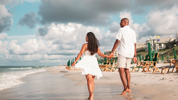 Un couple se promène sur une plage, main dans la main, avec des chaises et des parasols en arrière-plan.