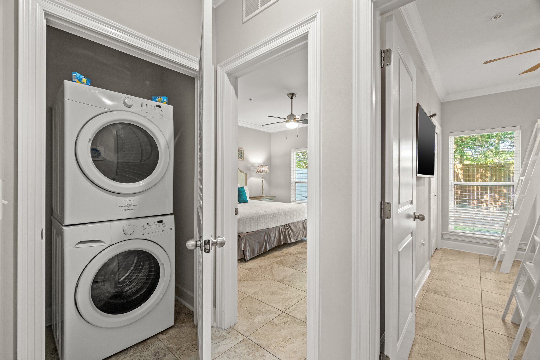 A laundry area with stacked washers and dryers, a bedroom with a bed and a ceiling fan, and a view of a backyard through a window.