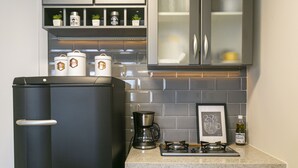 Una cocina con un refrigerador negro, una cafetera, una estufa a gas, y una alacena con frascos y un cuadro enmarcado.