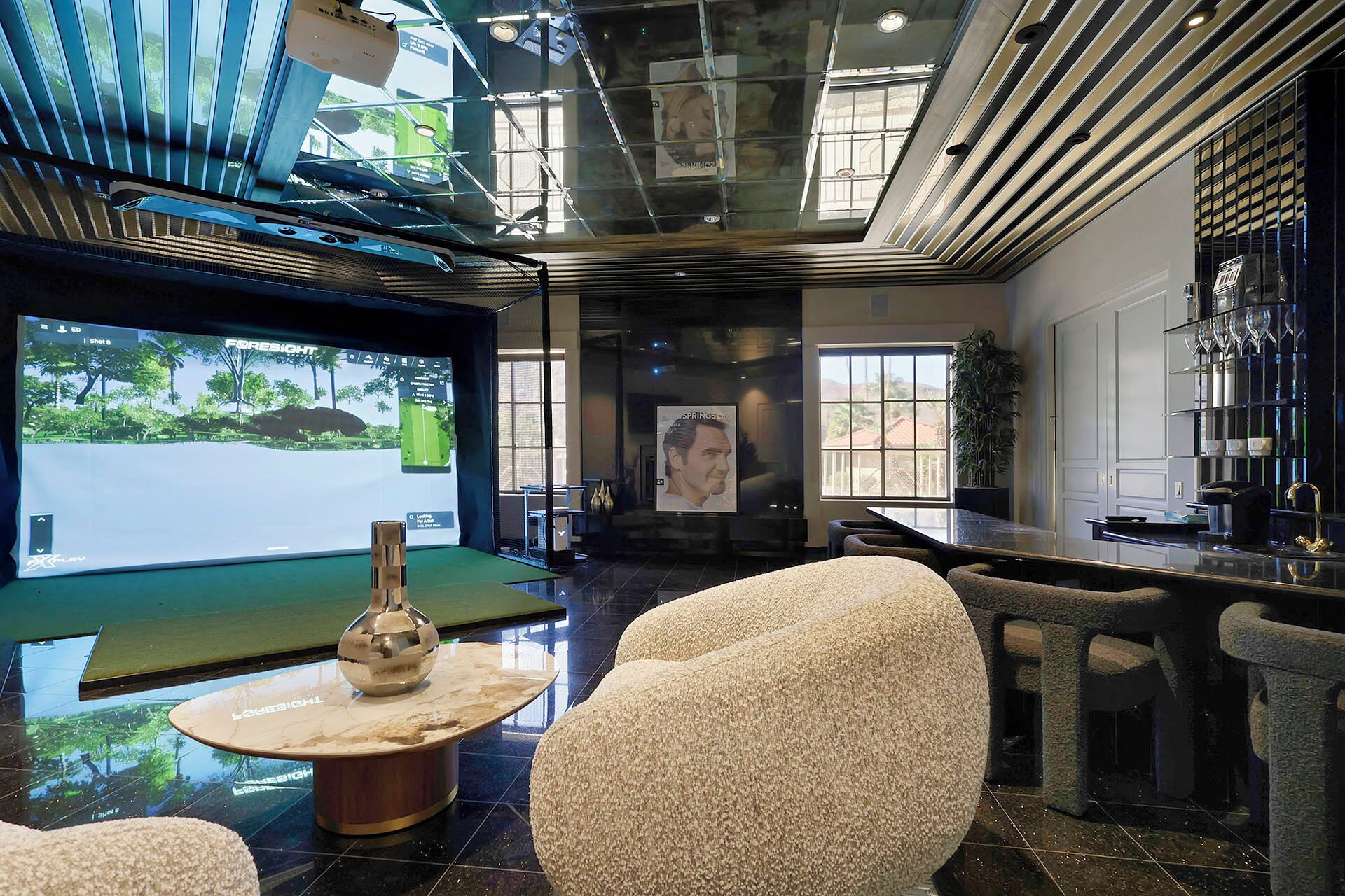 Un intérieur moderne avec un grand écran diffusant des images d’un parcours de golf, un coin bar avec des tabourets et un espace salon agrémenté d’un canapé à texture intéressante.