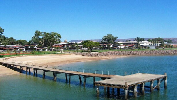 A coastal area with a wooden pier extending into calm waters, surrounded by a sandy beach and a row of buildings in the background.