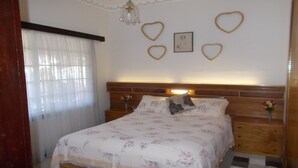 A bedroom with a wooden headboard, floral bedding, heart-shaped wall decorations, and a bedside table with a vase of flowers.