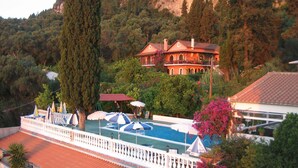 A pool area with umbrellas, a large tree, and a building with a red roof. - Romantic sea view Studio for two (Corfu)