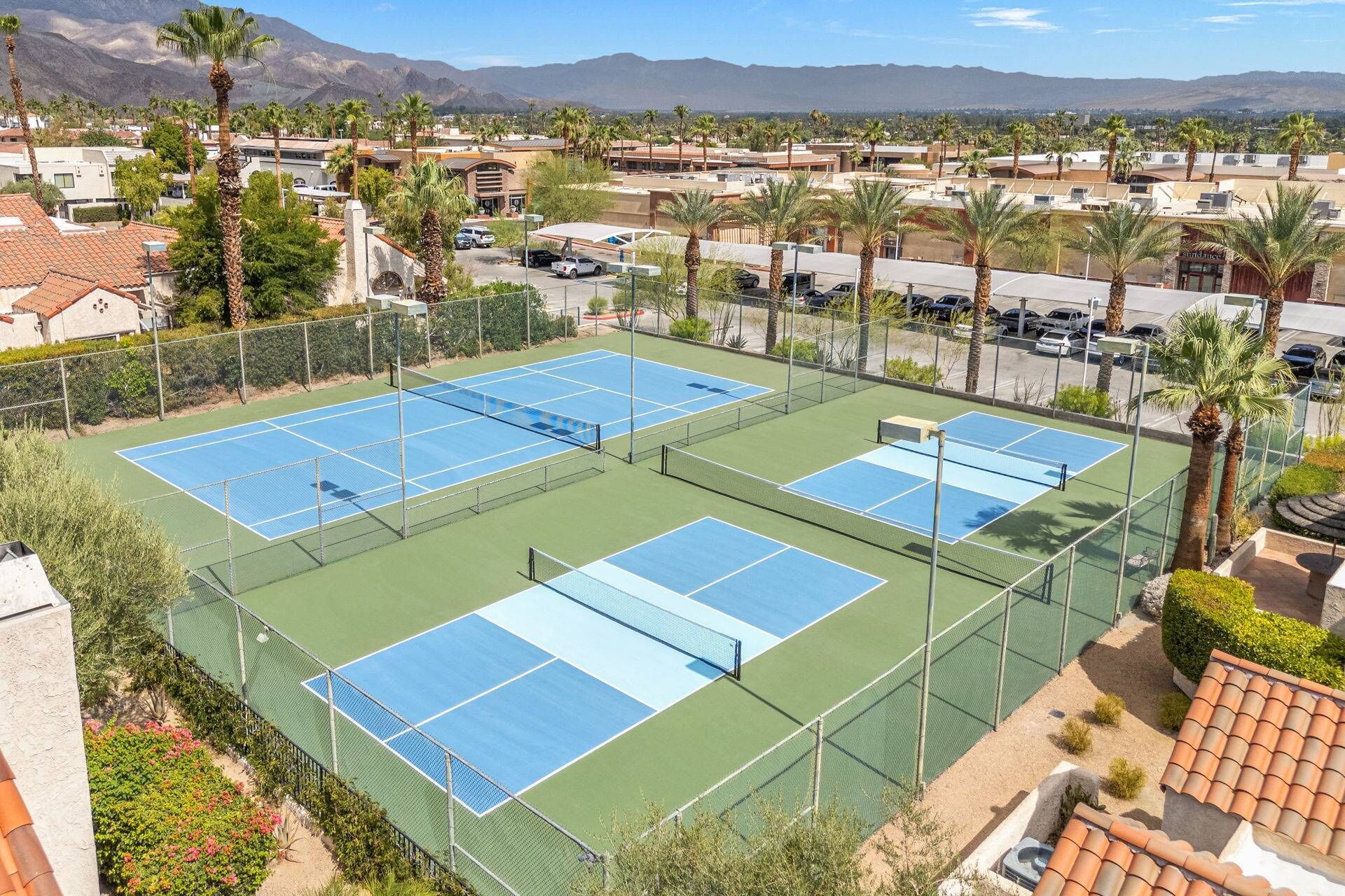 A tennis court complex with multiple courts, surrounded by a fence, palm trees, and a mountainous backdrop.