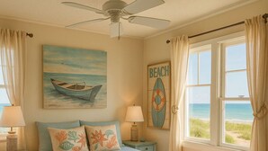 Ein Schlafzimmer mit Bett, Deckenventilator, einem Bild eines Bootes, einem Strandschild und Blick durch das Fenster auf den Strand.