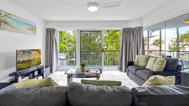Un salon moderne avec un canapé gris, une table basse et un téléviseur posé sur un meuble. Il y a un balcon d’où l’on peut admirer la verdure et un immeuble.