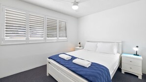 Une chambre d’hôtel avec un lit simple, des murs blancs, un ventilateur de plafond et une fenêtre avec des stores.