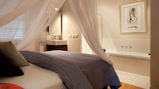 A bedroom with a bed, a bedside table, a nightstand, a window with blinds, a bathroom with a sink and bathtub, and a framed artwork on the wall. - Circa Red London Daylesford ~ Town Centre! (Daylesford)