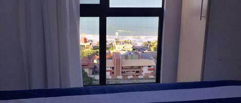 A bed with striped bedding and two towels with palm tree designs, a window with a view of a beach and buildings, and a white curtain.