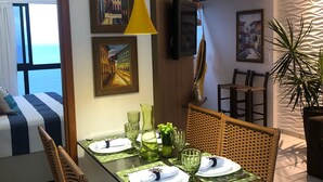 A dining area with wicker chairs, a glass table, and a yellow pendant light. There are framed artworks on the walls and a potted plant on a shelf. - Flat with spectacular view of PONTA NEGRA BEACH, 55 MB Wi-Fi, garage (Natal)
